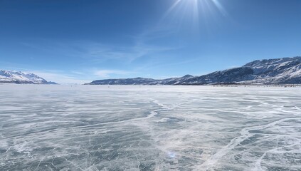 Obraz premium Icy lake surface with visible fissures overlooking a snowy shoreline and frosted mountain range on a bright, chilly winter afternoon.