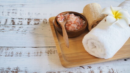 Spa composition with white rolled towel decorated with frangipani flower, Himalayan pink salt in wooden bowl with spoon, and loofah on wooden tray over rustic white wooden background