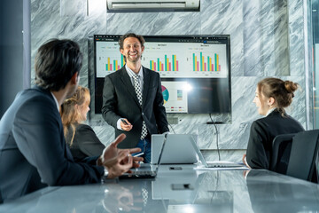 Smiling caucasian businessman leading corporate meeting in modern office room with multiracial colleagues sitting around table discussing business performance using chart presentation