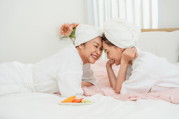 Asian senior woman lying on bed smiling closely with teenage daughter during home spa day both wearing white bathrobes towels wrapped on heads creating joyful beauty bonding moment