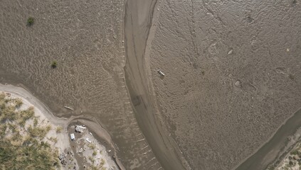 Overhead perspective of the tidal mudflats revealing winding water channels among grassy dunes and sandy shores, coastal island in the distance, abstract textures of water and sand