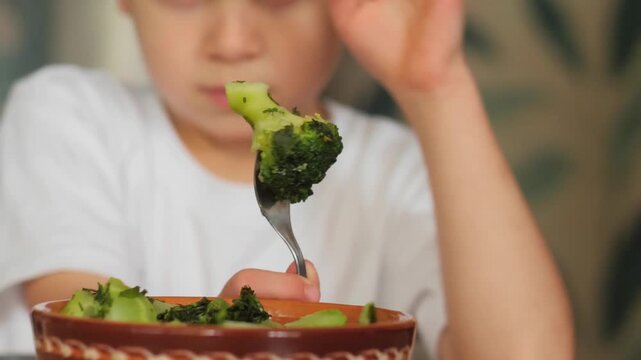 A boy looking skeptically at broccoli - A young boy holds up a fork with a single floret of broccoli and looks at it with a skeptical, unsure expression like he hate broccoli