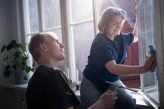 Young man holding squeegee while girlfriend cleaning window with cloth at home
