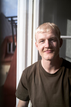 Portrait of smiling blond young man sitting on window at home