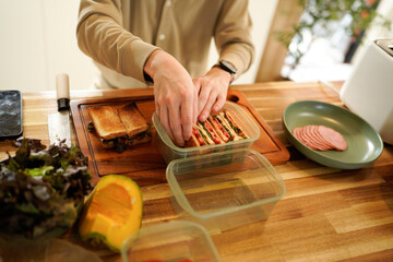 A man is preparing sandwiches in a kitchen. He is using a knife to cut the bread and is placing the sandwiches in plastic containers