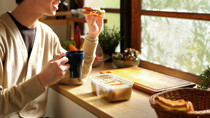 A man is eating a sandwich and drinking coffee Near the window in the morning. Scene is relaxed and casual