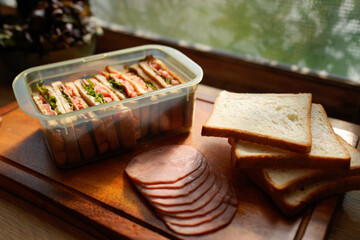 A tray of sandwiches and bread sits on a wooden cutting board Near a window with natural sunlight. The sandwiches are in a plastic container and the bread is sliced.