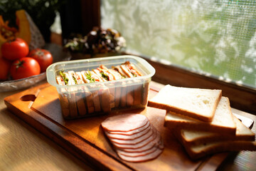 A tray of sandwiches and bread sits on a wooden cutting board Near a window with natural sunlight. The sandwiches are in a plastic container and the bread is sliced.