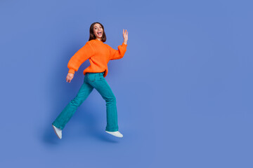 Young cheerful woman in an orange sweater and blue jeans dancing joyfully in front of a solid...