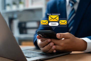 Businessman holding smartphone receives new email message alert while sitting at desk with open laptop. Represents modern digital communication, connectivity, and real-time email marketing workflow.