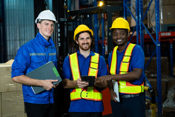 Group of three multicultural warehouse workers discussing operations, two men holding tablet and clipboard while wearing hard hats and safety vests inside industrial storage facility
