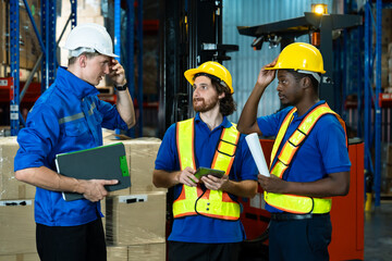 Multicultural warehouse team of three male adults discussing ideas during warehouse operation, one holding clipboard, another holding rolled documents, standing near forklift in storage area