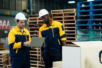 Asian and african adult male workers standing in warehouse with laptop, checking inventory status beside loaded boxes and pallets, representing multicultural work collaboration