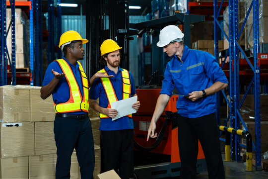 Multiracial male warehouse workers discussing task with supervisor using tablet among stacked cardboard boxes in distribution center background showing collaboration in logistics workplace