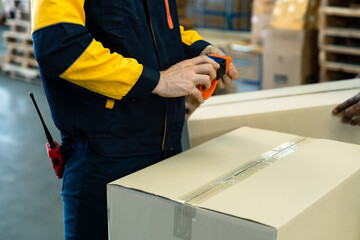 Male warehouse worker sealing cardboard box using packing tape for secure shipping during product handling process in storage area with stacked boxes and pallets, inside logistics warehouse.