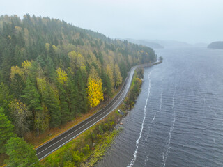 Drone view of an asphalt road hugging the shoreline of a large, dark lake, with a dense forest showing bright yellow and green autumn colors