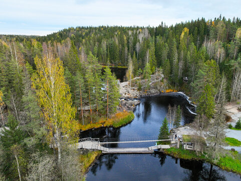 Aerial view of a long wooden footbridge crossing a dark river surrounded by a dense northern forest in autumn colors