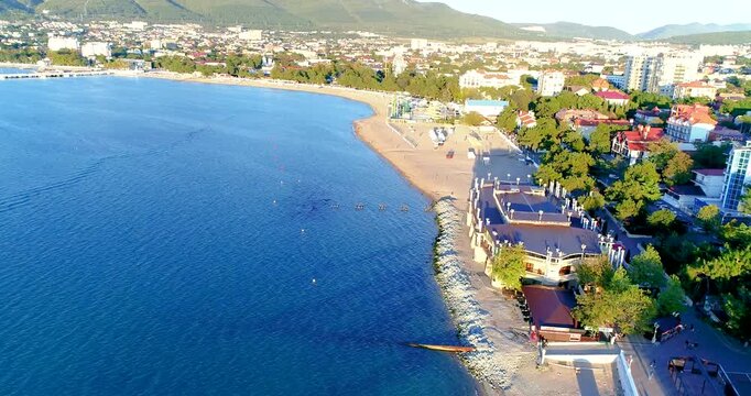 Flight over central beach resort of Gelendzhik. Promenade with balustrade runs along beach. Umbrellas and sunbeds on beach. Blue sea. Caucasus Mountains in background.