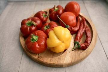 Vibrant red and yellow peppers, chili peppers, and tomatoes arranged on a round wooden board. A classic still life representing the healthy bounty of the autumn harvest season. 
