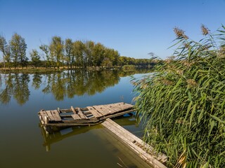a dilapidated wooden fisherman's platform on the bank of a flat river, the mirror-like surface of which reflects the trees on a cloudless sunny summer day