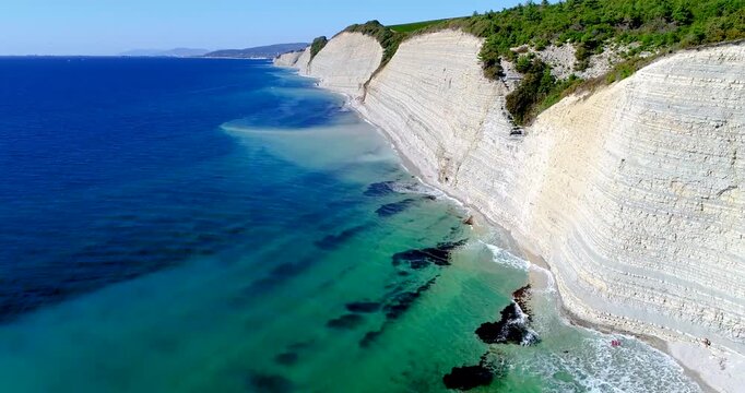 Flight over Cherno Sea coast. High layered rock with small pebble beach below. Trees grow above, Beautiful blue sea. Surf. Sunny weather