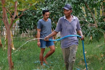 A father moves across his farmland with flowing hose water to find more plants to irrigate, while his young son follows closely behind him in the village landscape.