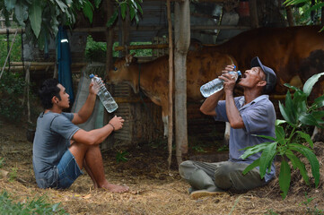 After working on their farmland, a father and son rest by the cow shed, sharing bottled water and enjoying a short break in the afternoon sunlight.