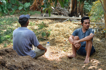 A farmer father and his son sit on hay beside dried cow dung, observing the calm farmland in front of the cattle shed during the afternoon.