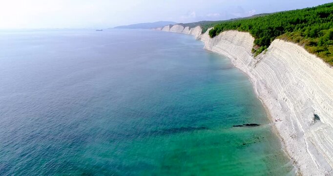 Flight over Cherno Sea coast. High layered rock with small pebble beach below. Trees grow above, Beautiful blue sea. Surf. Sunny weather