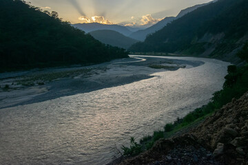 Teesta river flowing through Himalayan mountains, sunlight coming through clouds in the background, setting sun behind mountain peaks. Sikkim, India.