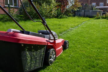 A lawn mower stands on a green lawn in the yard of a house with well-groomed bushes and a garden.