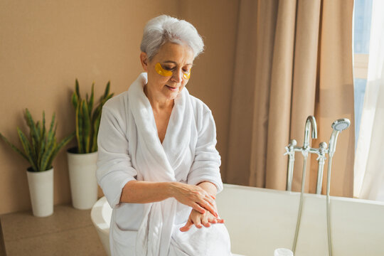 Mature adult old woman applying moisturizing body lotion on hands. Senior lady rubbing nourishing anti age cream doing morning daily skincare routine in bathroom. Beauty treatment spa body skin care - Powered by Adobe