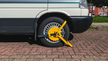 Naklejka premium Detail shot of metal clamp securing motorhome wheel beside curb, bright daylight shows strict parking control