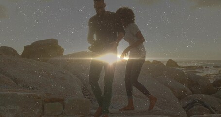 Strolling couple barefoot traversing rocky shoreline at sunset, with lens flare and waves