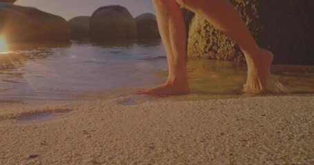 Stepping barefoot man wearing trunks into shallow water on beach, rounded boulders, rock formation