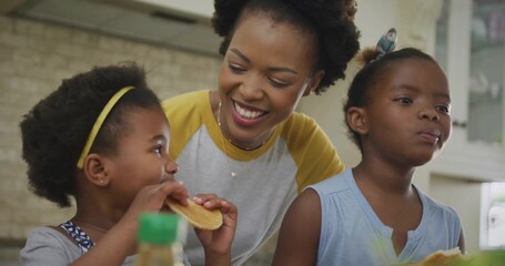 Leaning mother and daughters eating pancakes at kitchen counter with syrup bottle and pancake stack