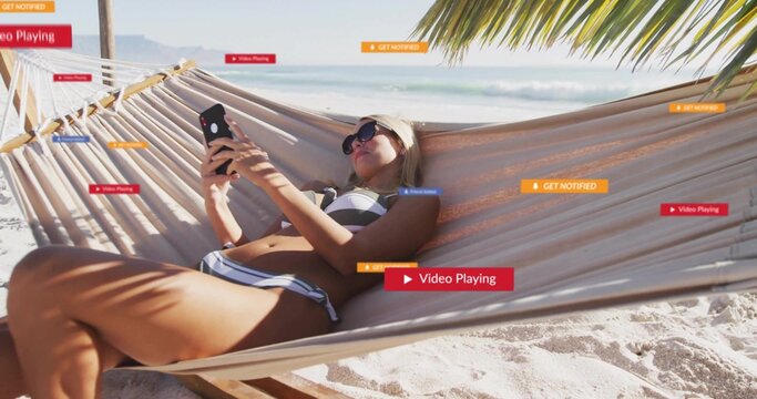 Reclining woman wearing swimsuit, sunglasses and headband, holding smartphone in hammock at beach