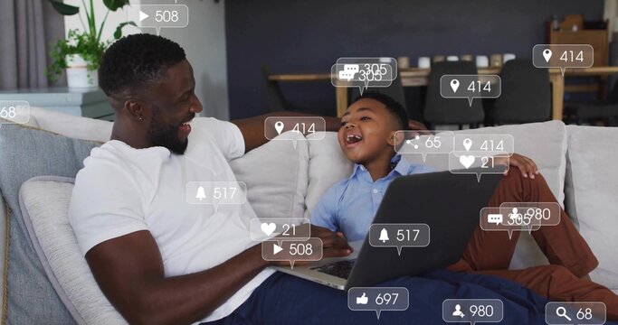 Leaning father and son sharing laptop on sectional sofa in modern living room, with potted plant