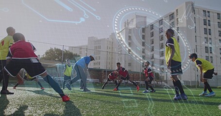 Coach in tracksuit, cap guiding boys stretching on turf soccer field, with goal net, fence, benches