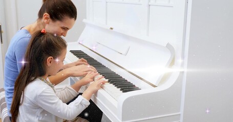 Sitting mother and daughter pressing keys on white upright piano at home, with bench and sparkles