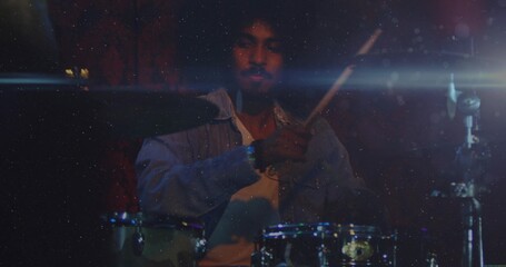 Playing drummer wearing light denim shirt over white T-shirt in dimly lit stage, with drum kit