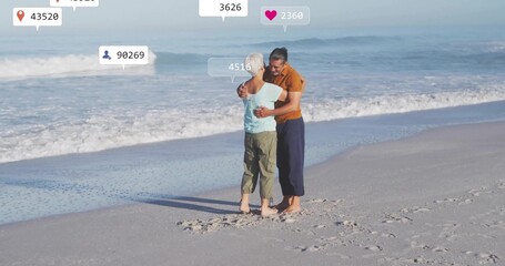 Embracing senior couple standing on sandy beach at water edge, with floating digital overlays
