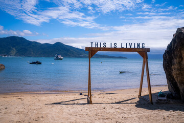 A wooden beach frame with the words “This Is Living” stands on a sandy shore, overlooking boats on the ocean with lush mountains in the background under a partly cloudy sky.