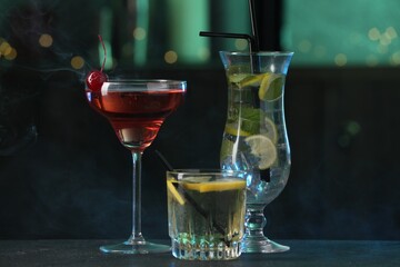 Glasses of tasty cocktails on dark table against blurred background with smoke, bokeh effect
