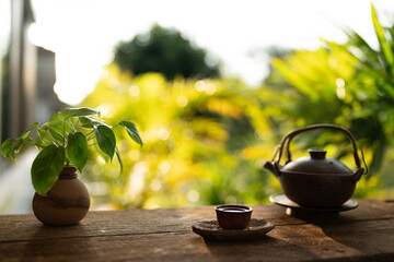 Tea cup and black kettle tea pot on wooden table with pothos plant, with balcony view under sunlight