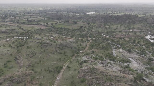 Aerial shot of two white cars driving along a dirt path through rocky hills near Masuda, Rajasthan, surrounded by green Aravalli vegetation, small ponds, and water bodies under a cloudy monsoon sky.