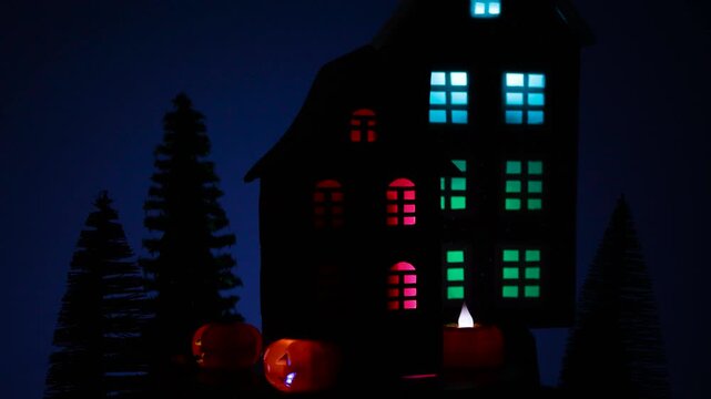A Halloween house surrounded by glowing pumpkins and candles rotates among Christmas trees against the backdrop of a large black house