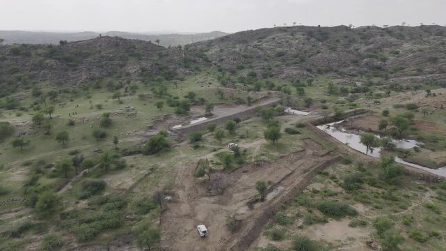 Aerial shot of two white cars driving along a dirt path through rocky hills near Masuda, Rajasthan, surrounded by green Aravalli vegetation, small ponds, and water bodies under a cloudy monsoon sky.