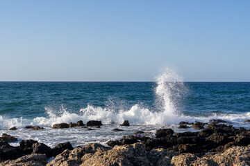 Powerful sea waves crashing against the rocky coastline of Crete, Greece, under bright blue sky. The dynamic movement of the water and spray captures the raw beauty and energy of the Mediterranean Sea