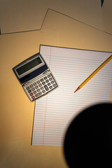 a handheld calculator, a pencil, and notebook paper under the light of a desk lamp, moody lighting 